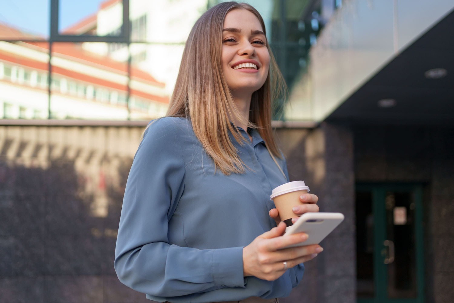 Mulher sorri ao usar o celular na rua, em frente a um prédio público, para acompanhar um processo digital da prefeitura